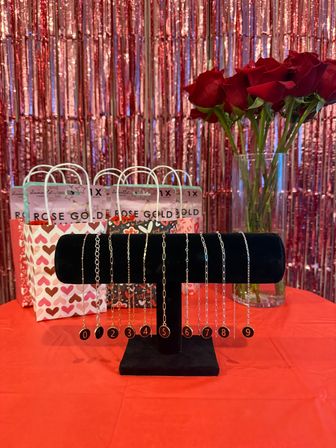 Valentine's Day retail display with numbered charm bracelets on a black velvet stand, bouquet of red roses in a vase, heart-patterned gift bags and shimmering rose-gold fringe backdrop on a red table.