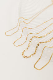 Close-up of gleaming gold-tone layered necklaces with mixed chain styles, including paperclip and beaded links, draped over a white ribbed tray — minimalist jewelry flatlay