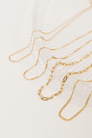 Close-up of gleaming gold-tone layered necklaces with mixed chain styles, including paperclip and beaded links, draped over a white ribbed tray — minimalist jewelry flatlay