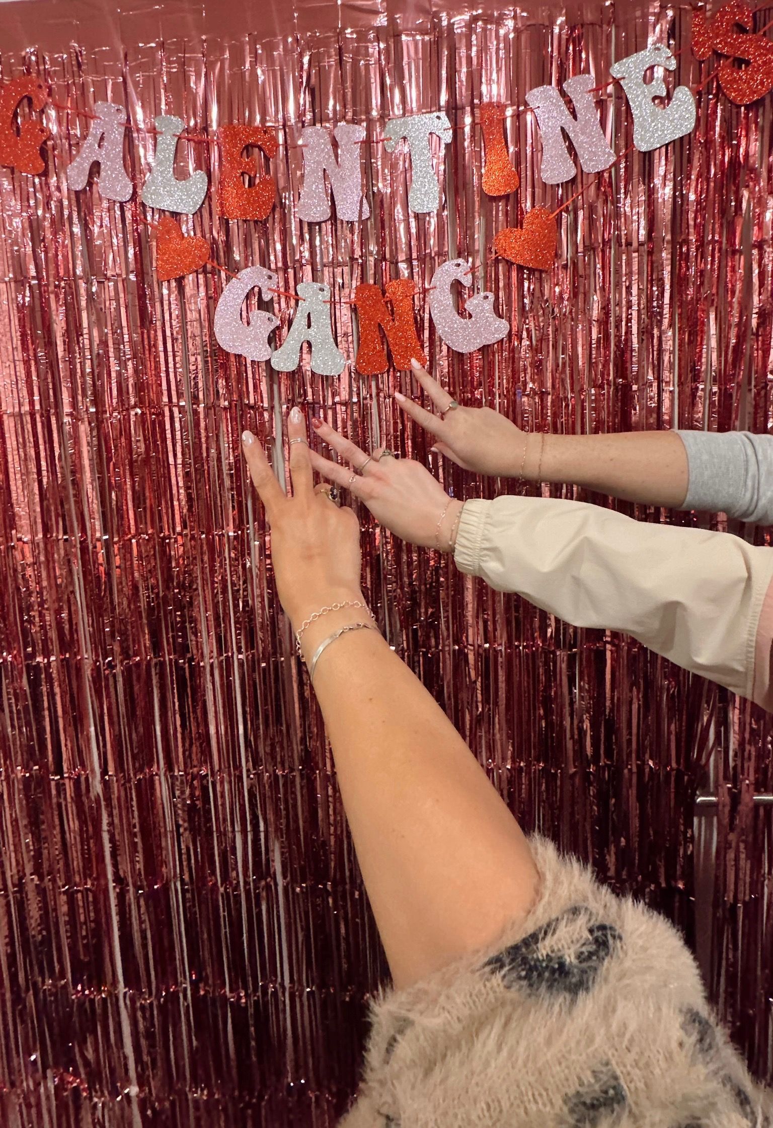 Pink tinsel Valentine's photo booth backdrop with glitter "Valentine Gang" banner and three hands posing peace signs, showing rings and bracelets.