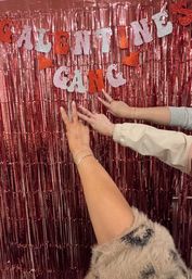 Pink tinsel Valentine's photo booth backdrop with glitter "Valentine Gang" banner and three hands posing peace signs, showing rings and bracelets.