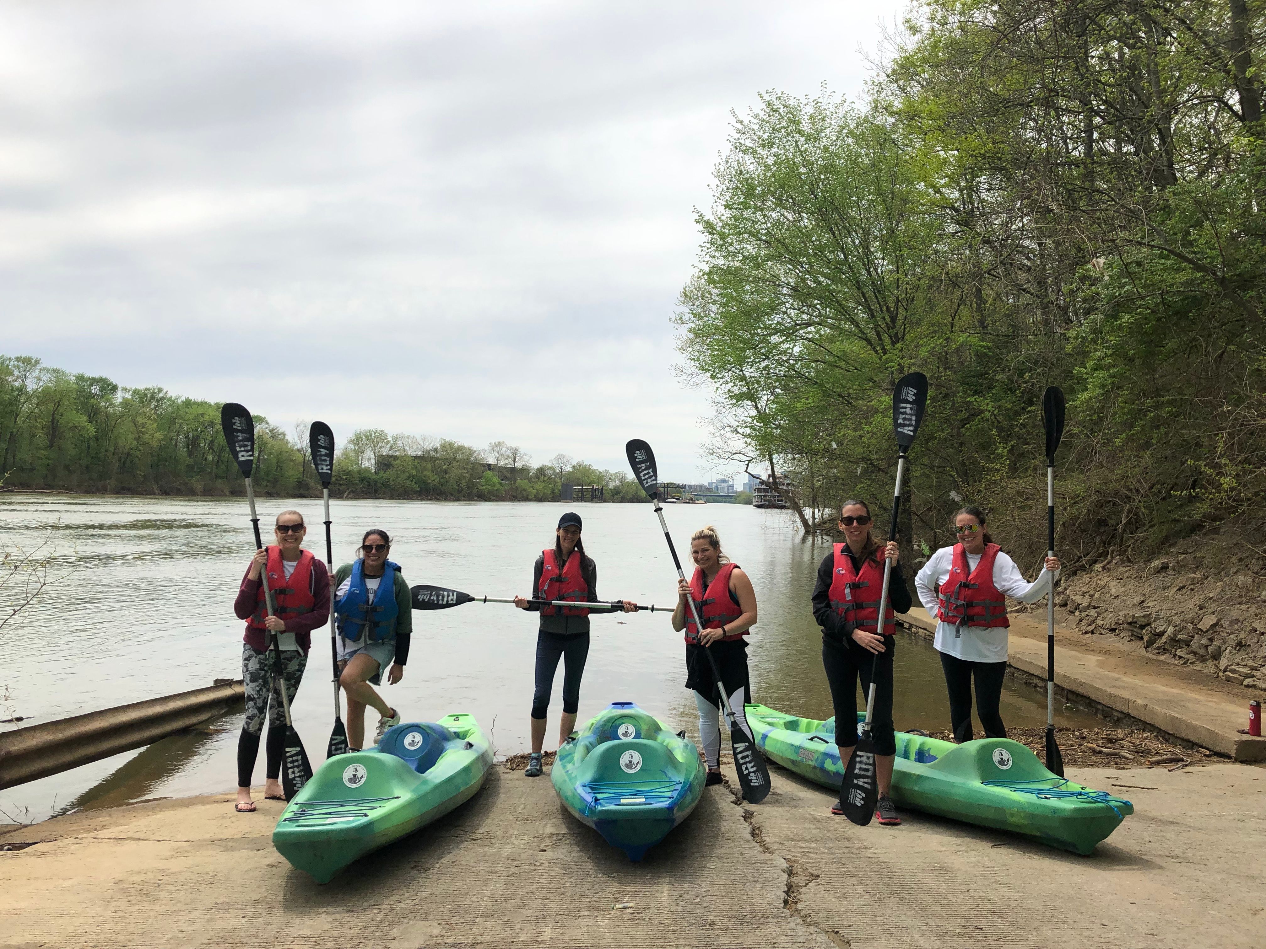 Six paddlers in life jackets stand at a concrete river launch holding paddles beside three green kayaks, with a calm, tree-lined river and overcast sky in the background.