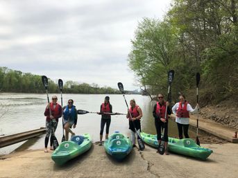 Six paddlers in life jackets stand at a concrete river launch holding paddles beside three green kayaks, with a calm, tree-lined river and overcast sky in the background.