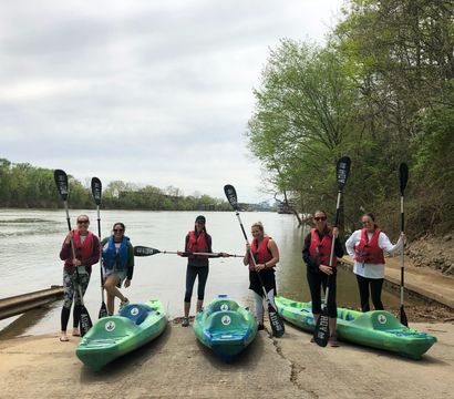 Six paddlers in life jackets stand at a concrete river launch holding paddles beside three green kayaks, with a calm, tree-lined river and overcast sky in the background.