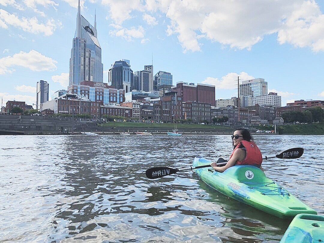 Kayaker in a bright green kayak paddling on the Cumberland River with downtown Nashville skyline and a distinctive pointed skyscraper under a sunny blue sky.