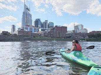 Kayaker in a bright green kayak paddling on the Cumberland River with downtown Nashville skyline and a distinctive pointed skyscraper under a sunny blue sky.
