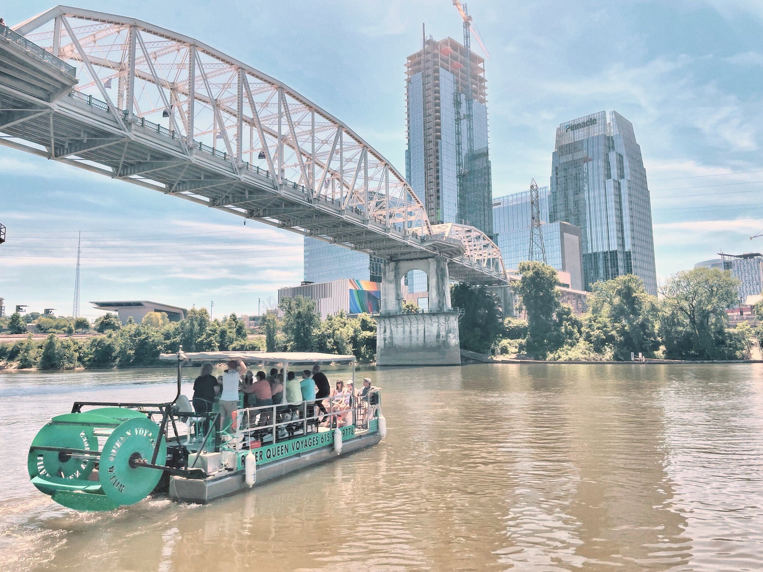 Paddlewheel tour boat with passengers cruising the Cumberland River under a steel arch bridge with downtown Nashville glass skyscrapers and green riverbank on a sunny day