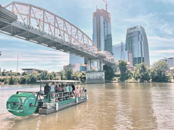 Paddlewheel tour boat with passengers cruising the Cumberland River under a steel arch bridge with downtown Nashville glass skyscrapers and green riverbank on a sunny day