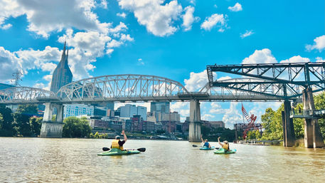 Three kayakers paddling on the Cumberland River under steel truss bridges with the Nashville downtown skyline and bright blue sky