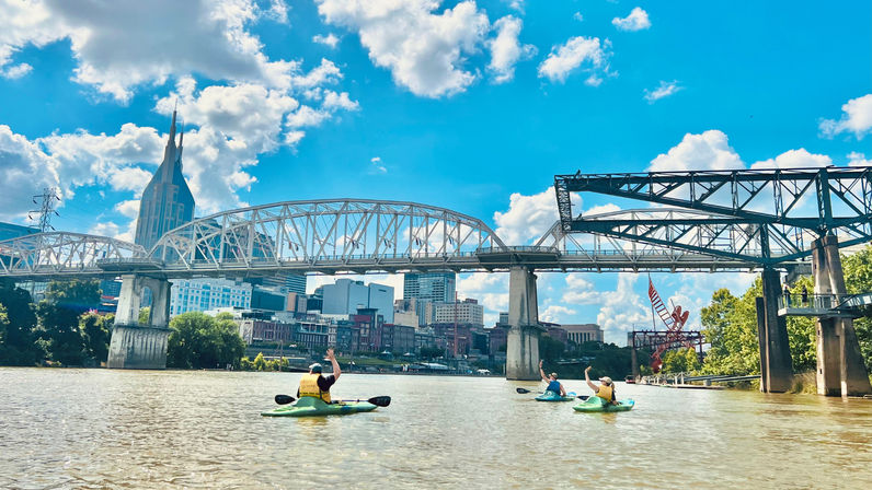 Three kayakers paddling on the Cumberland River under steel truss bridges with the Nashville downtown skyline and bright blue sky