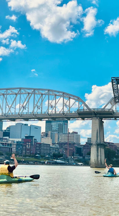 Three kayakers paddling on the Cumberland River under steel truss bridges with the Nashville downtown skyline and bright blue sky