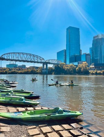 Sunlit urban riverfront with green kayaks lined on a stone ramp, paddlers gliding on calm water beneath a steel truss bridge and modern glass downtown skyscrapers.