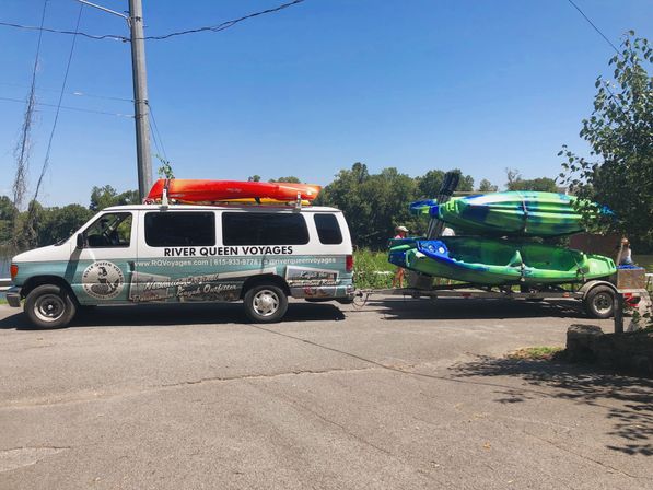 White transport van towing a trailer stacked with green and blue kayaks and an orange kayak on the roof, parked at a sunny riverfront launch with trees and clear blue sky.