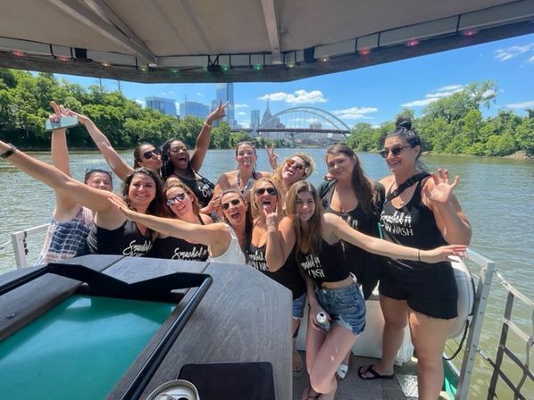 Cheerful group of women in matching black tank tops posing and laughing on a sunny riverboat cruise, with a city skyline and arched bridge framed by green riverbanks in the background.