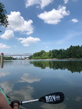 View from a kayak on a glassy river reflecting fluffy clouds, with treelined banks, a steel railroad bridge and distant city skyline under a sunny blue summer sky