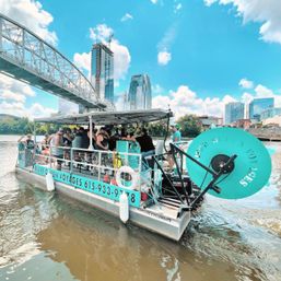 Turquoise pedal-powered party boat full of passengers cruising a sunny downtown riverfront under a steel bridge with glass high-rises