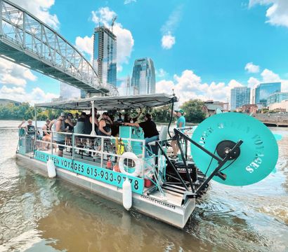 Turquoise pedal-powered party boat full of passengers cruising a sunny downtown riverfront under a steel bridge with glass high-rises