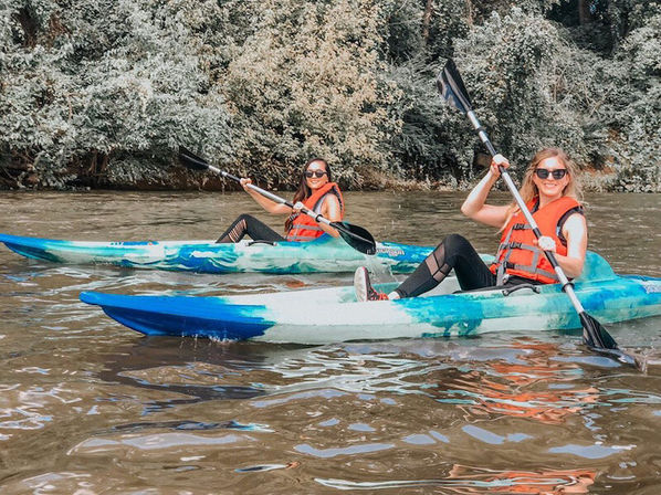 Two people in orange life jackets paddling blue-and-white kayaks on a calm, tree-lined river — relaxed outdoor kayaking adventure.