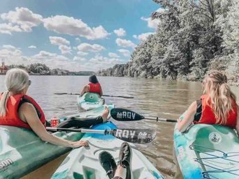 Three kayakers paddling on a calm river under a bright blue sky with fluffy clouds and tree-lined shore, view from one kayak showing feet and paddle in the foreground — summer outdoor water adventure.