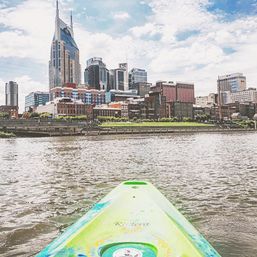 Green kayak bow on the Cumberland River pointing toward downtown Nashville skyline under a bright blue sky.