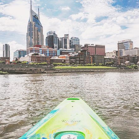 Green kayak bow on the Cumberland River pointing toward downtown Nashville skyline under a bright blue sky.