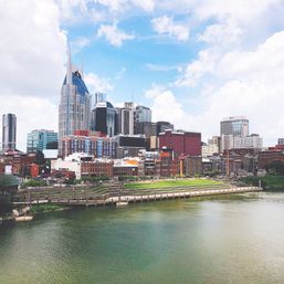 Sunny Nashville skyline along the Cumberland River, downtown skyscrapers rising above historic brick riverfront and a green riverwalk.