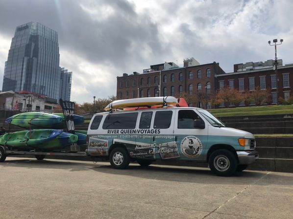 Kayak rental van and trailer loaded with colorful kayaks parked on a downtown riverfront steps, with brick warehouse buildings and a glass skyscraper under a cloudy sky