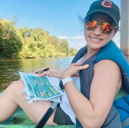 Smiling kayaker in a cap and mirrored sunglasses wearing a turquoise life jacket, holding a pen and laminated papers while paddling on a calm tree-lined river under a blue summer sky.