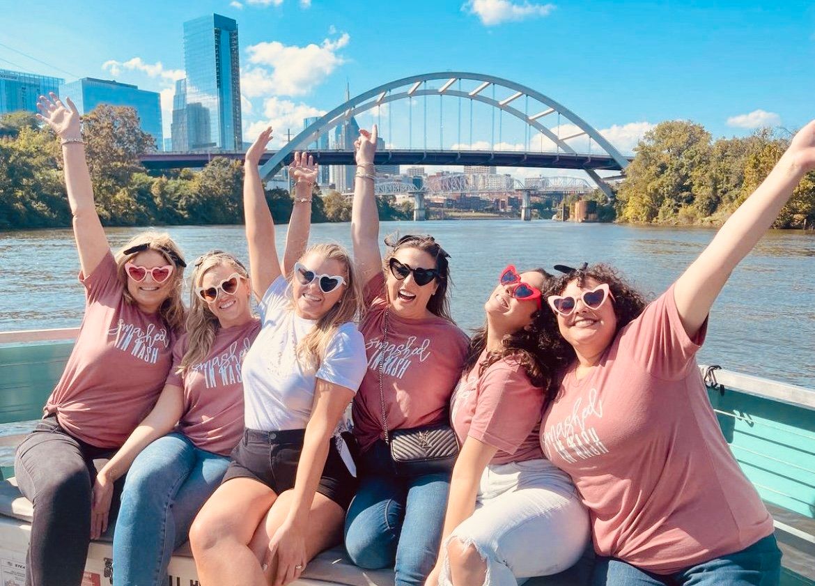 Six friends in matching pink shirts and heart-shaped sunglasses cheering on a sunny riverboat cruise with an arched bridge and downtown skyline in the background.