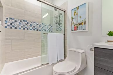 Bright modern residential bathroom with glass sliding shower, blue patterned tile accent, white tub and one-piece toilet, palm tree wall art.