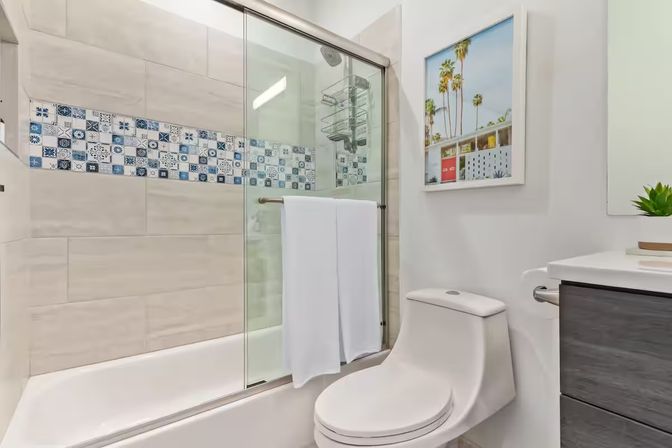 Bright modern residential bathroom with glass sliding shower, blue patterned tile accent, white tub and one-piece toilet, palm tree wall art.
