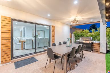 Covered modern backyard patio at dusk with a long rectangular dining table and eight black chairs, stainless steel gas grill, sliding glass doors revealing a bright indoor dining room, overhead light fixture and hedge privacy screening