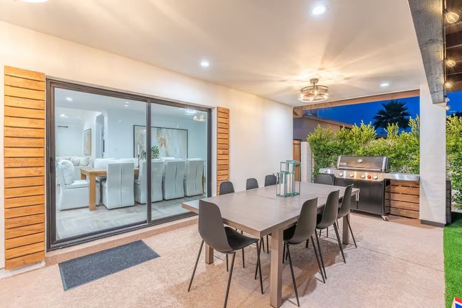 Covered modern backyard patio at dusk with a long rectangular dining table and eight black chairs, stainless steel gas grill, sliding glass doors revealing a bright indoor dining room, overhead light fixture and hedge privacy screening
