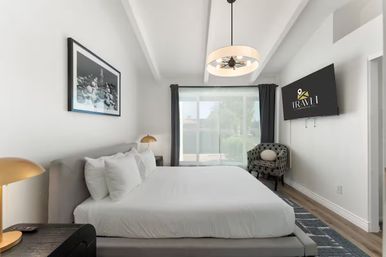 Bright modern hotel bedroom with king bed in white linens, gold bedside lamps, patterned accent chair, wall-mounted TV, and large sliding glass door letting in natural light