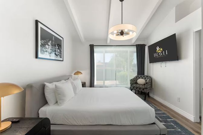 Bright modern hotel bedroom with king bed in white linens, gold bedside lamps, patterned accent chair, wall-mounted TV, and large sliding glass door letting in natural light