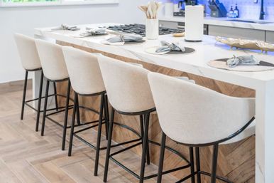 Row of beige upholstered bar stools with black metal legs at a white quartz kitchen island with chevron wood base and place settings — modern bright open-concept kitchen seating.