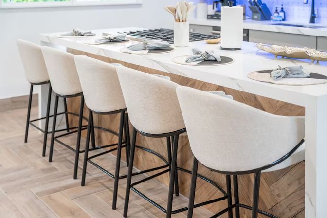 Row of beige upholstered bar stools with black metal legs at a white quartz kitchen island with chevron wood base and place settings — modern bright open-concept kitchen seating.