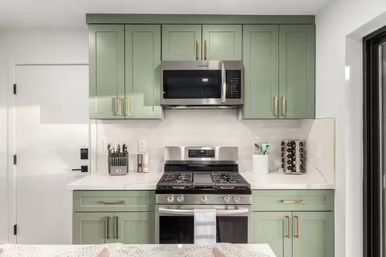 Bright modern kitchen with cheery sage-green shaker cabinets, brass hardware, stainless steel microwave and gas range, white quartz countertops, knife block and spice rack.