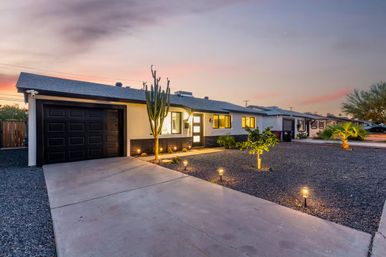 Southwestern single-story ranch home at dusk with illuminated pathway lights, black garage door, gravel desert landscaping, tall cactus and small palm