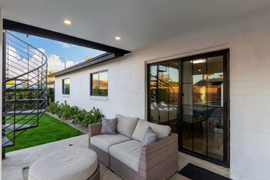 Modern covered backyard patio with wicker loveseat and ottoman, sliding glass door reflecting a black spiral metal staircase, manicured lawn and garden bed along a white brick house.