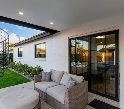 Modern covered backyard patio with wicker loveseat and ottoman, sliding glass door reflecting a black spiral metal staircase, manicured lawn and garden bed along a white brick house.