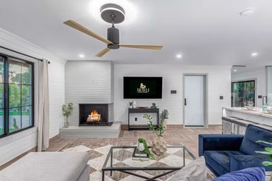 Bright modern open-concept living room with wood-blade ceiling fan, lit fireplace set in white brick, wall-mounted TV, large window with garden view, blue velvet sofa and glass coffee table with potted plants near a kitchen island.