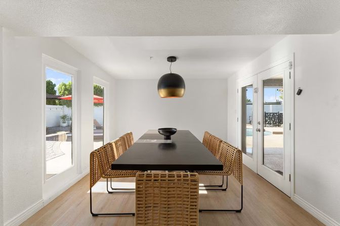 Sunlit modern dining room with a long matte-black table, woven rattan chairs, gold-accent pendant light, and glass doors overlooking a backyard pool.