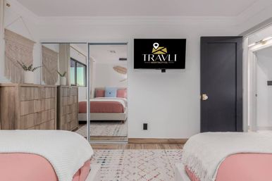 Bright modern boho guest bedroom with two pink-accented beds, wooden dressers, macramé wall hangings, mirrored closet doors, wall-mounted TV, and a patterned area rug.