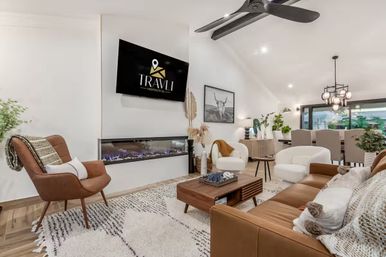 Bright contemporary open-plan living room and dining area with tan leather sofa, white armchairs and mid-century lounge chair, wooden coffee table, built-in linear electric fireplace under a wall-mounted TV, patterned rug, ceiling fan, pendant light and indoor plants.
