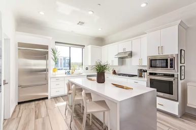 Bright modern open-concept white kitchen with waterfall island and three bar stools, stainless-steel refrigerator and double oven, gas cooktop, potted plant centerpiece, window view and light wood-look tile floor.