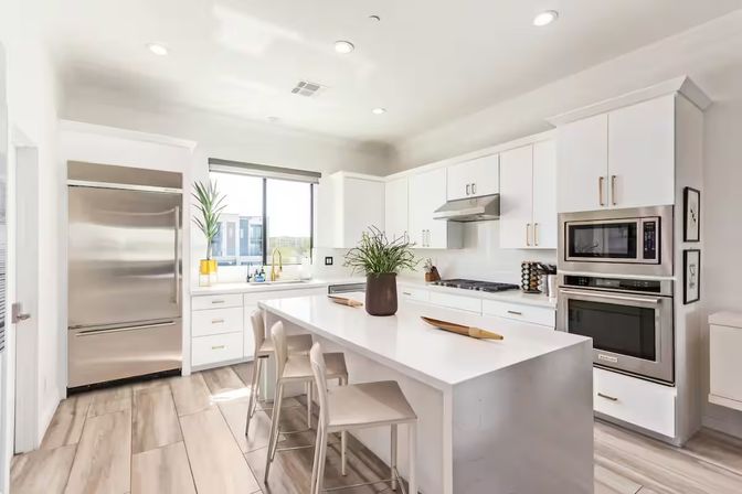 Bright modern open-concept white kitchen with waterfall island and three bar stools, stainless-steel refrigerator and double oven, gas cooktop, potted plant centerpiece, window view and light wood-look tile floor.