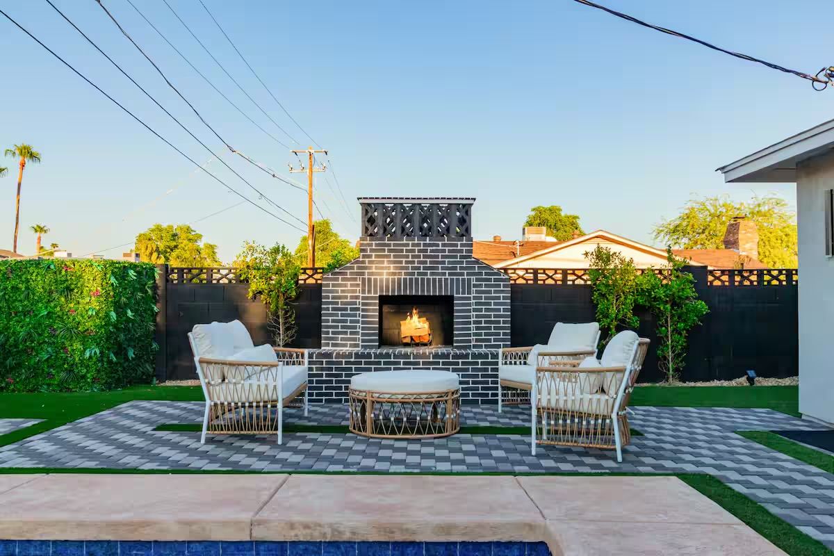 Cozy suburban backyard patio with black-and-white brick outdoor fireplace, small fire, rattan wicker lounge chairs and ottoman on a patterned paver terrace beside a swimming pool