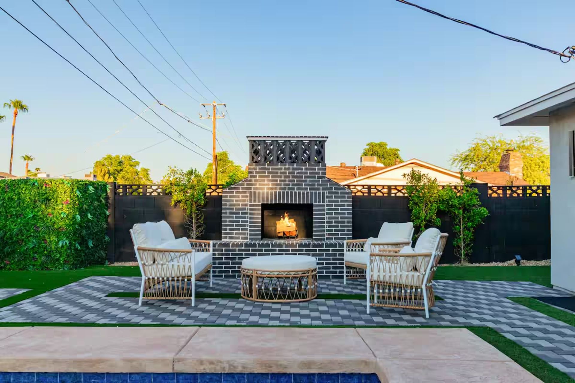 Cozy suburban backyard patio with black-and-white brick outdoor fireplace, small fire, rattan wicker lounge chairs and ottoman on a patterned paver terrace beside a swimming pool