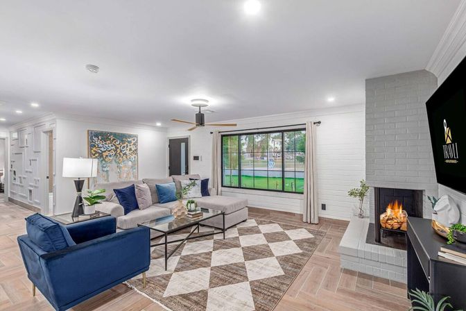 Cozy contemporary living room with gray sectional and navy armchair, glass coffee table on a geometric rug, lit fireplace, ceiling fan, and large black-framed window overlooking a green yard.